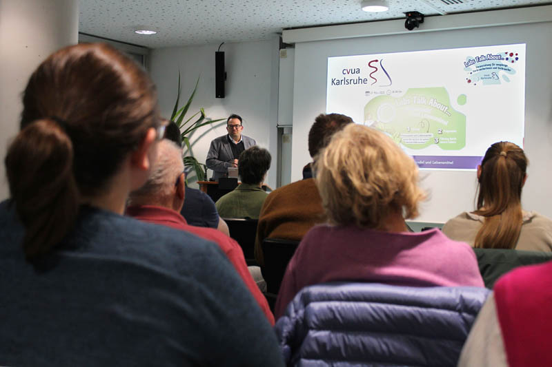 Im Hintergrund ist der Amtsleiter des CVUA Karlsruhe, Dr. Walch, zu sehen, der vor einem interessierten Publikum im Vordergrund einen Vortrag über die Arbeit des CVUA Karlsruhe und die Herausforderungen der Lebensmittelüberwachung vor dem Hintergrund des Klimawandels hält.